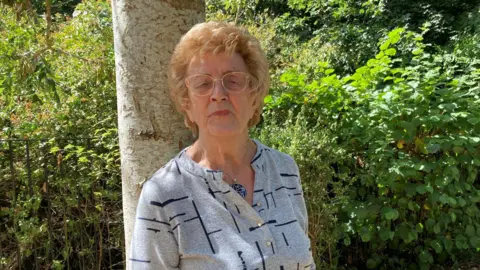 BBC/Naj Modak Ann Bentley wearing glasses stands outdoors beside a large tree trunk. The person is wearing a light grey long-sleeved top with a dark linear pattern and a pendant necklace. The setting is a leafy, green area with dense vegetation, bushes, and trees in the background. Sunlight filters through the foliage.