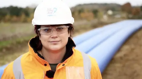 John Fairhall/BBC Regan Harris smiles at the camera in front of three blue water pipes. She is wearing a white coloured hard hat and an orange hi-vis coat. She is also wearing clear safety glasses. Her dark hair has been tucked into the hard hat.