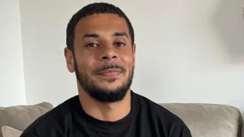 Jordan Chiedozie sitting on a sofa wearing a black t-shirt, with a plain background