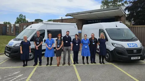 NHS Humber Health Partnership NHS staff – some wearing medical uniforms – lined up between the white vans, which have NHS logos on the front.