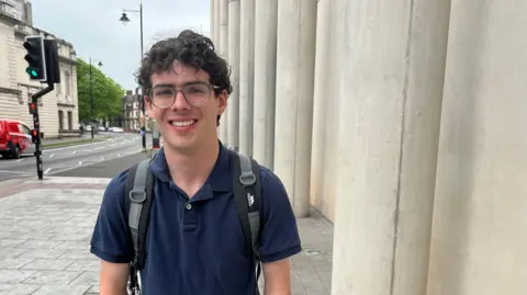A young man with dark curl hair and glasses. He is wearing a navy blue polo shirt and a grey and black backpack and is smiling and looking at the camera. Behind him are the pillars of a university building on one side, and a road with traffic lights on the other.