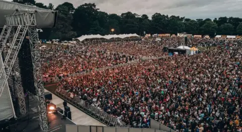 Bedford Park Concerts An outdoor festival audience, pictured from one side of the stage, showing the mixing desk area and tents/foodstalls around the arena perimeter.