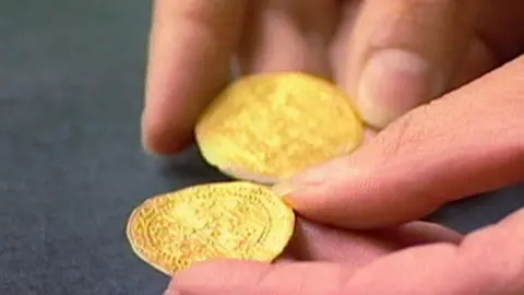 A close-up image shows a hand holding two small gold-coloured coins or medallions with intricate decorative patterns stamped into their surfaces. The objects are resting on a dark tabletop, with the foreground coin in sharp focus while the second coin and fingers behind it appear slightly blurred.