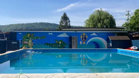 A picture of an outdoor swimming pool at a primary school. Behind it is a rectangular one-storey building with a colourful mural painted on the side, depicting crocodiles, fluffy clouds, a wave and the school's crest logo. In the distance there is a large hill covered with thick forest. The sky is blue with white wispy clouds. 