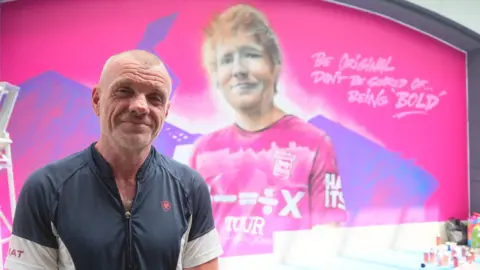 Keith Hopewell a man who is looking directly at the camera and smiling. He is standing inside a shopping centre in front of a bright pink wall which has a mural of Ed Sheeran on. 
