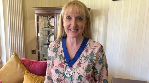 Claire Whiteway: she has long blonde hair and wears a salmon-pink floral dress, with her captain's medal around her neck on a blue ribbon.