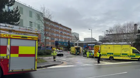 A fire engine and several ambulances are parked up on roads around Southampton General Hospital. A police officer can be seen standing at the end of the road leading to the hospital. Several other emergency service personnel can be seen walking around the scene in hi-viz clothing. 