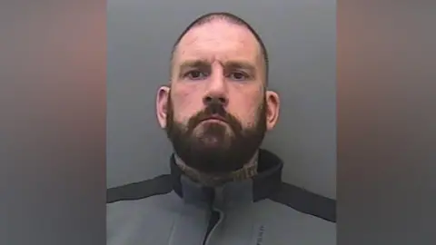 Gary Parkinson looking at the camera. He has short black hair and a full beard. He is wearing a black and grey tracksuit jumper. 