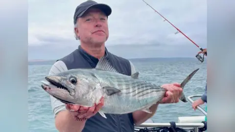 David Kiddy is wearing a navy blue waistcoat and shorts and baseball cap and is holding a large silvery-grey fish. His hands are supporting its head and its tail.