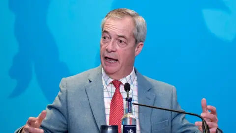 Nigel Farage sat at a desk in front of a blue backdrop, gesticulating with his hands. He is wearing a grey suit, red tie with white spots and a chequered shirt.