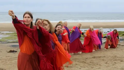 A line of women in highly coloured clothes holding hands and pulling hard at an imaginary rope on a beach.