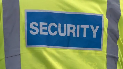 A close up of a yellow hi-viz vest with grey stripes on the left and right. In the middle is a blue rectangle with the words security written in capital letters.