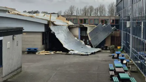 A secondary school building with a section of the roof blown off and lying in what looks like a playground area