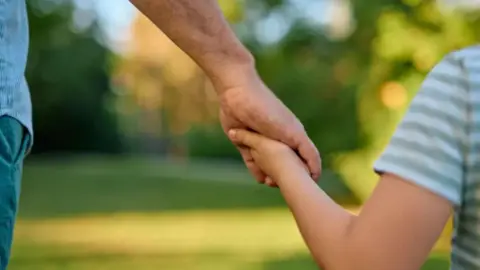 A father and son holding hands as they are walk through a green area. 
