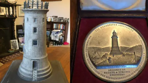 Jennifer Kloester The model of the Plymouth breakwater lighthouse on the left. It is grey and has steps at the bottom which alternate. There are two larger windows at the centre of the lighthouse and three smaller ones a the top. On the left is a picture of the medallion which has the writing 'The Breakwater Lighthouse' across the top and at has William Stuart's name on the outside. It also has a picture of the completed lighthouse and is in a red box.