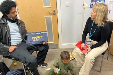 Two-year old Omer plays with toys on the floor as his father, who is seated, speaks to the community nurse, who is also seated, in an office at Walthamstow Family Hub