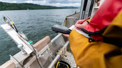A scientist - who wears yellow overalls and a red life jacket - records some figures from a machine and writes them down with a pencil on paper. They are standing on the side of a boat which is sailing on Windermere.