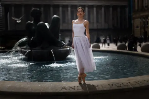 Clive Booth A dancer wearing a white top and tulle skirt, and ballet slippers stands on her toes on the edge of a water fountain.
