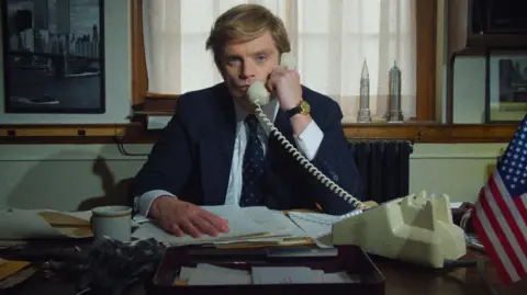 Studio Canal Sebastian Stan as Donald Trump in The Apprentice - he is on a white landline phone while sitting at a desk strewn with business papers. He is dressed in a navy suit and looks very serious.