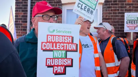 Men standing on a picket line. In the foreground is a man who can only partially be seen, he is wearing a blue jacket, a red baseball cap on his head and a pair of glasses. He is holding up a sign in front of his body which says, RUBBISH COLLECTION NOT RUBBISH PAY. Behind him are men wearing orange fluorescent hi-vis vests and are holding placards.