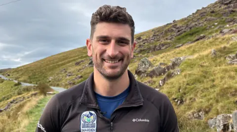 Alun Jones of Eryri National Park Authority smiling at the camera, standing on one of the paths up Yr Wyddfa
