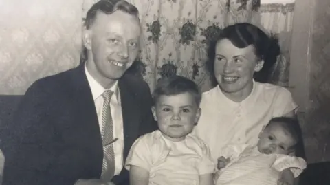 William Cruickshank A black and white picture of William Cruickshank, centre, as a child with his family. His father, William Crockett is on the left of the picture, wearing a dark suit, light shirt and striped tie. William is in the middle and is wearing a light T-shirt shirt. His mother, wearing a light blouse is on the right, holding William's sister, who is a baby and wearing a light top. They are sitting in front of curtains with a floral pattern.