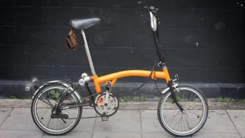 An orange Brompton folding bicycle is on a pavement next to a wall.
