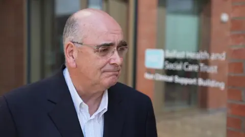 Mr Mulgrew is looking to the right of the frame. He is wearing thin framed glasses and has a plain expression. He is bald and wearing a navy suit jacket, and a white shirt with no tie and the top button undone. 