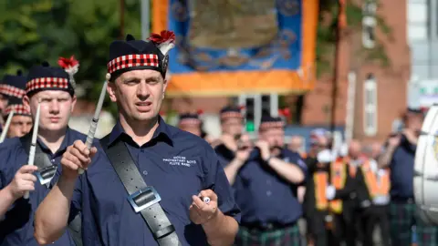 Members of a band parading down the street wearing navy short sleeved shirts and tartan bordered hats with a red flower. The sun is shining on the band members. 