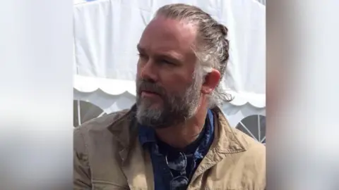 A man with a grey beard and hair tied back looking to the side. He is wearing a beige jacket and a blue shirt with sunglasses hanging on.