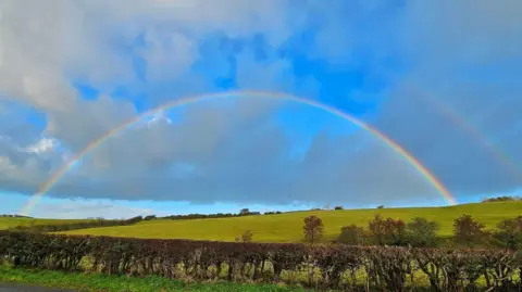 A rainbow over some green fields, a fainter rainbow can be seen above it. The sky is blue with white and grey clouds. There is a hedge in the foreground and trees in the field.