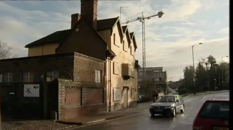A side view of a boarded-up pub building. It is over three floors. A car is passing on the road outside and a crane is rising in the distance. The road is wet, the sky clouded. 