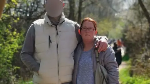 Facebook Marta Bednsizic with short brown hair and brown glasses wearing a blue top and a gray coat with a gray bag, standing next to a person whose face is blurred on a row -bound footpath by trees.