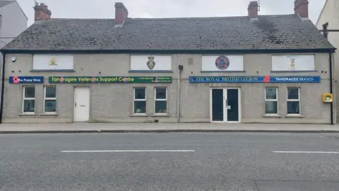 A detached building on a street that has two sets of doors.  The doors on the left  are underneath a sign saying 'The Poppy Shop' and 'Tandragee Veterans Support Centre'.  The door on the right is underneath a sign saying 'The Royal British Legion Tandragee Branch'