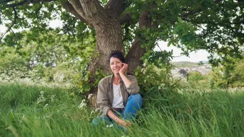 Natalie Fee sits under an oak tree in a grassy meadow 
