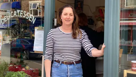 A woman stands in the doorway of a small delicatessen shop, holding the doorframe with one hand. The person is wearing a black‑and‑white striped long‑sleeve shirt, blue jeans, and a belt. Behind the person, the shop interior is visible with shelves, signage, and another customer inside.