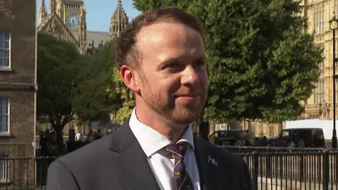 North Cornwall MP Ben Maguire stood outside Westminster in the sunshine. He is looking away from the camera with a smile. He is wearing a black suit and a white shirt with a Cornish flag pin badge. 