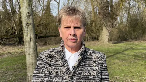 BBC Woman, with blonde hair, wearing a black and white jacket and white shirt, looking expressionless, with a trees and grass behind her.