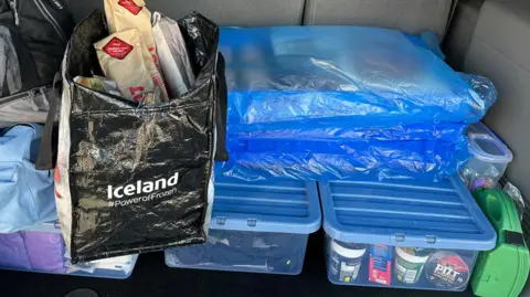 The interior of a van boot with blue lidded plastic crates of food and clothing, crates of baked goods wrapped in blue plastic and a bag full of sandwiches are piled on top
