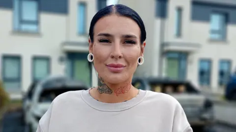 A woman, with black hair tied back, standing in front of a row of houses. She is wearing a cream coloured jumper and has white earrings. She has tattoos on her neck. 