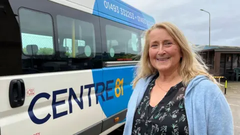 Alison Holmes, wearing a patterned black top, and blue cardigan, has long blonde hair. She is smiling and standing next to one of the charity's fleet of minibuses which is adapted for people with physical and learning disabilities.