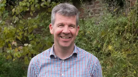A headshot of Paul Learoyd, chief executive of Lincolnshire Wildlife Trust. He is stood in front of a green bush and is smiling at the camera. He has short grey hair and is wearing a red, blue and white checked shirt.