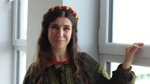 A woman with long brown hair stands by a window in the daytime, smiling into the camera. She wears a dark green traditional Ukrainian blouse with embroidery on the front and sleeves, and a headband decorated with small red and yellow flowers.