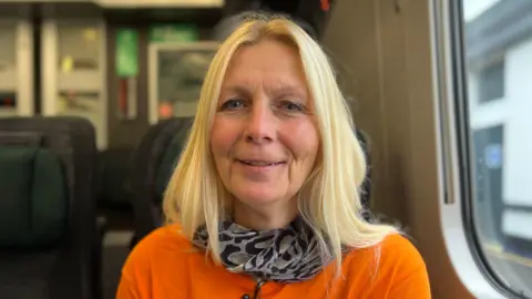 Julie Braithwaite sitting on a train. She has blonde hair and is wearing an orange shirt. She is smiling.