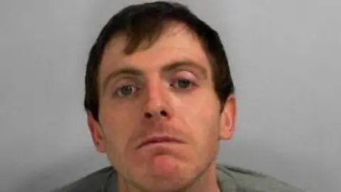 A mugshot of a man who is looking slightly down towards the camera. He is wearing a grey jumper and has brown hair, which is partly combed over his forehead.