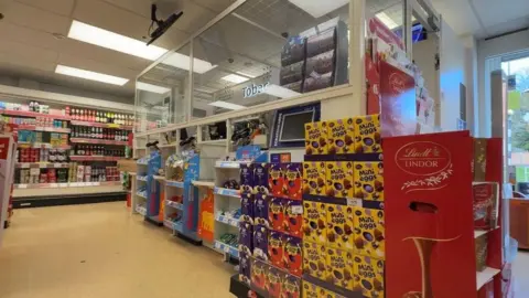Glass screens wrap around a kiosk till in a shop. Tobacco products can be seen inside the screen. Outside the screen are stacks of Easter eggs of various brands and colours. Shelves full of other produce can be seen in the background.