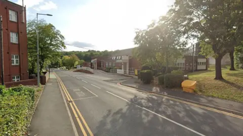 A road runs between two low-rise hospital buildings. There are double yellow lines on both sides and tree-lined grass verges. 