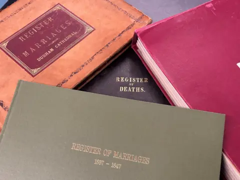 Four large record books in a messy pile on a desk. One is green and reads "register of marriages 1957-1847" on, another is old and orange with "register marriages Durham Cathedral" on the front, the third is black with "register of deaths" in gold letters and the final one is red.
