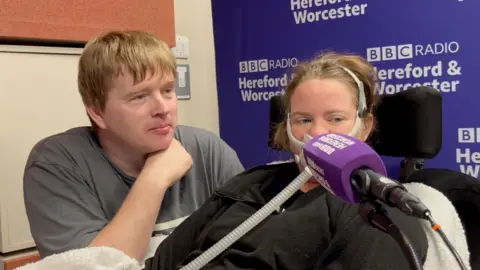 A man in a grey t-shirt next to a woman with motor neurone disease who has an oxygen tube attached to her face in a radio studio