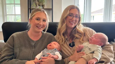 Abi holding her baby Freddie and Jessica with their baby Alfie. They are both smiling at the camera and sat next to eachother.